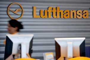 Eine Lufthansa-Mitarbeiterin geht am Flughafen in München an einem Logo der Fluggesellschaft vorbei., © dpa/Andreas Gebert