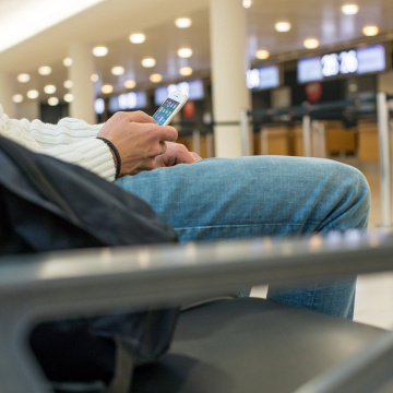 Ein Passagier sitzt mit dem Handy in der Hand auf einer Bank im Flughafen., © DPA/Christin Klose